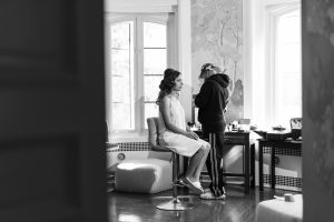 A woman in a white dress sits on a stool while a makeup artist stands in front of her, applying wedding makeup in a well-lit room with large windows and elegant decor.