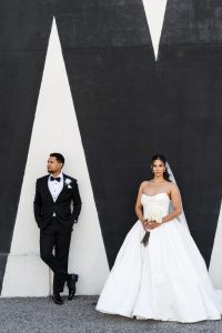 A bride in a white gown and a groom in a black tuxedo pose in front of a black-and-white geometric wall; the wedding photographer captures the groom leaning casually while the bride holds her bouquet and stands facing forward.