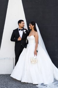 A bride in a strapless white gown and veil holds a bouquet of white roses, standing next to a groom in a black tuxedo. They smile at each other against a black and white geometric wall as the wedding photographer captures the moment.