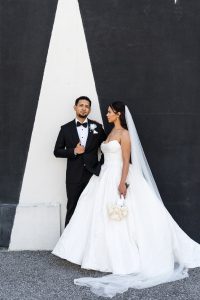 A groom in a black tuxedo and a bride in a strapless white gown with a long veil stand together against a geometric black and white wall, captured in striking documentary photography as the bride holding white flowers looks at the groom.