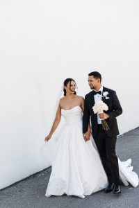 A bride in a strapless white gown and a groom in a black tuxedo walk hand in hand outside, smiling at each other. Captured by a Toronto photographer, the groom holds a bouquet of white flowers; the background is a plain white wall.