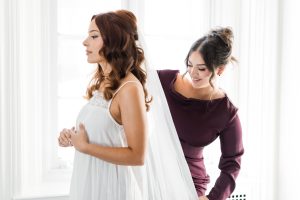 A bride in a white dress stands with her eyes closed as another woman in a long-sleeved burgundy dress adjusts her veil, captured by a wedding photographer in a bright Toronto room with large windows.
