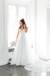 A bride in a white strapless wedding dress stands by a bright window, holding a bouquet and gently touching her hair. Captured in stunning photography, the airy room features white walls and dark wood floors.