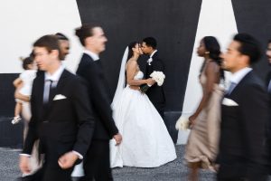 A bride and groom kiss while holding hands and a bouquet, wearing a white dress and black tuxedo, as their wedding photographer captures this documentary moment with guests in formal attire walking by in a blur.