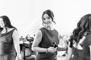 A woman in a formal dress smiles at the camera while holding a small object. Two other women in similar dresses stand nearby in a bright, indoor setting. This black and white image captures classic Toronto photography style.