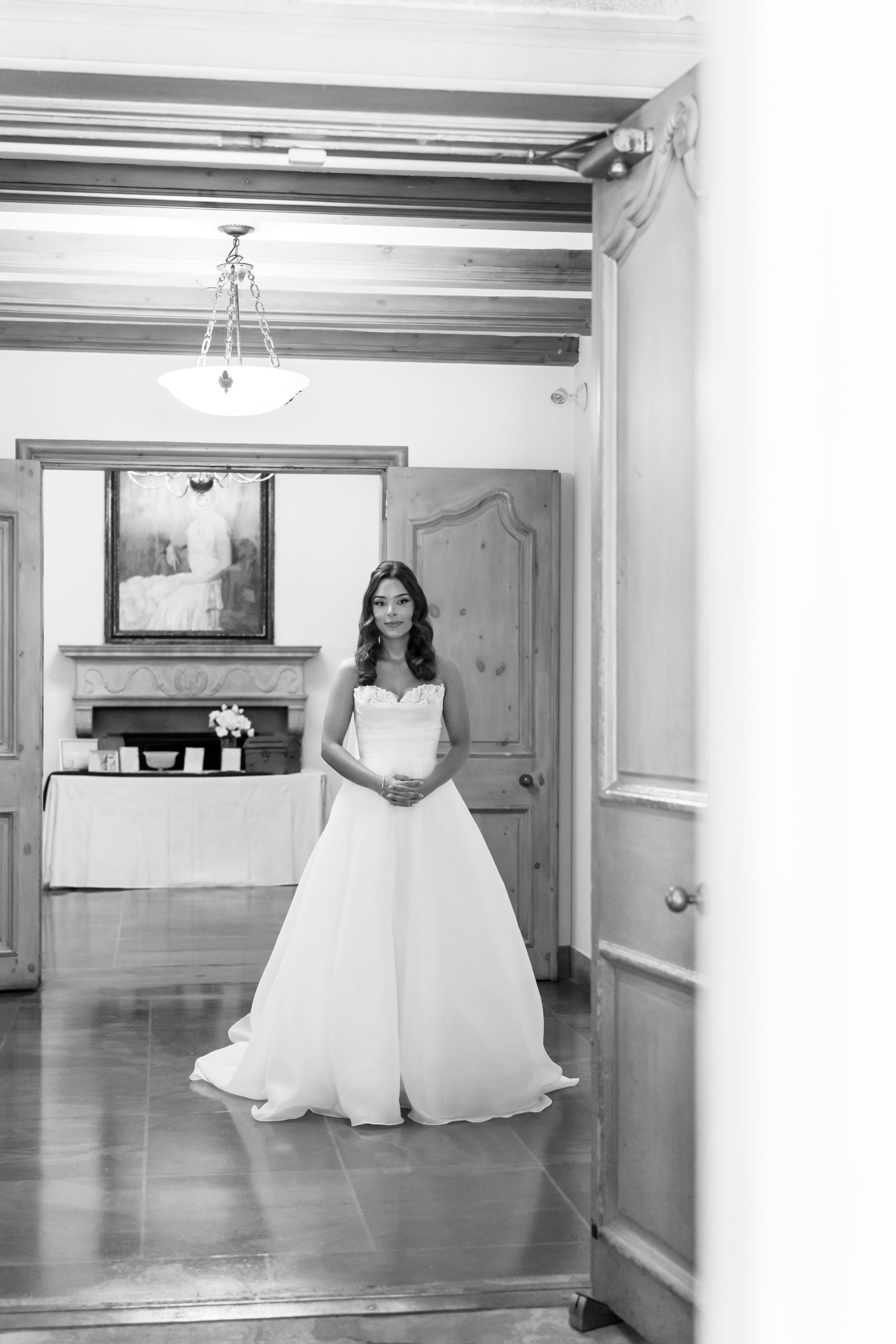 A bride in a strapless white gown stands calmly in the center of a room with wooden doors and a chandelier, captured in black and white. This Toronto wedding documentary moment frames her with open doors as she clasps her hands, looking ahead.