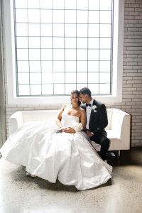 A bride in a white wedding dress and groom in a black tuxedo sit closely on a white couch, smiling and leaning toward each other, as natural light pours in through the window—capturing the perfect Toronto wedding photography moment.