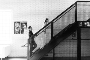 A bride and groom in wedding attire walk up a modern black staircase with a mesh railing against a white brick wall, passing posters labeled BRIDAL SUITES and BAR COATS—captured in a documentary style by their photographer.