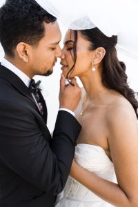 A groom in a black suit gently holds his bride’s chin as they lean in for a kiss under a white veil, captured with an editorial touch. The bride wears a strapless dress and pearl earrings.