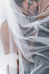 A bride and groom share an intimate moment under a sheer white veil, their faces close together as they gently touch and embrace. The soft fabric creates a dreamy, romantic atmosphere in this editorial wedding photograph.