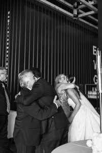 Black and white documentary photo of two couples embracing warmly at a wedding event; a bride and a woman hug on the right, while two men embrace on the left, all dressed formally, with onlookers in the background.