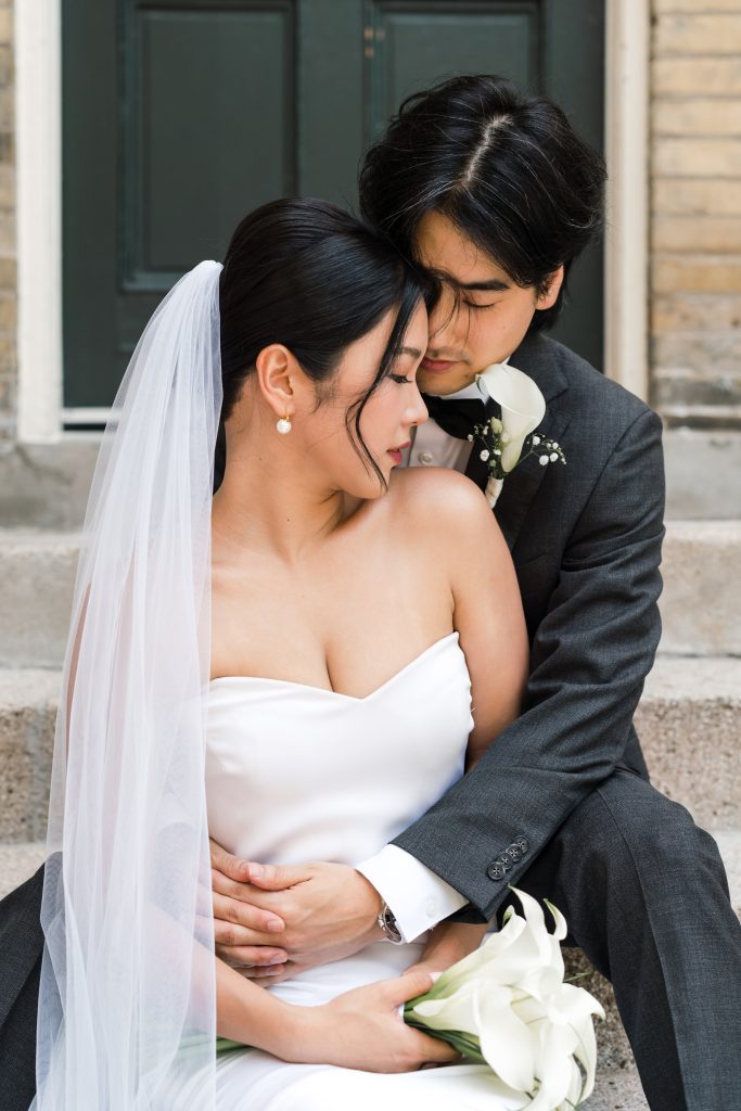 A bride and groom sit on outdoor steps in Toronto, embracing closely. The bride wears a white dress and veil, holding white flowers, while the groom in a dark suit hugs her from behind. Their wedding photography captures an intimate, serene moment.