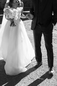 A black and white photography of a bride in a strapless gown holding a bouquet of roses, standing next to a man in a suit. They are holding hands and walking, with sunlight casting wedding shadows on the ground.