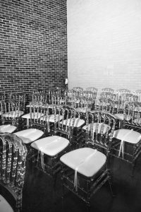 Rows of empty transparent chairs with white cushions are arranged in a room with dark and light brick walls, ready for a wedding ceremony—an inviting scene awaiting guests and the keen eye of a photographer to capture each moment.