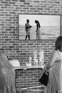 A woman in Toronto watches a TV mounted on a brick wall, displaying a black-and-white scene of a couple standing together by the ocean—perhaps from a wedding documentary. A table below holds glass containers and decorative items.