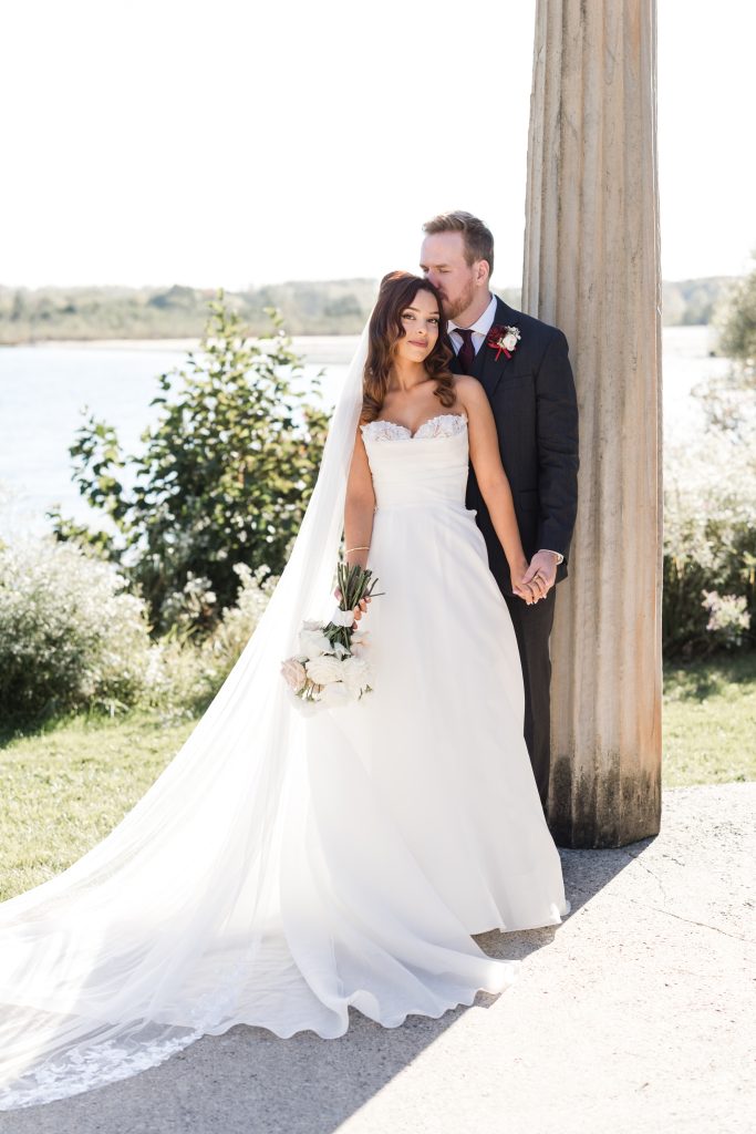 A bride in a white gown and long veil stands beside her groom in a dark suit by a column outdoors, holding a bouquet, with greenery and a lake in the background—a timeless editorial wedding scene.