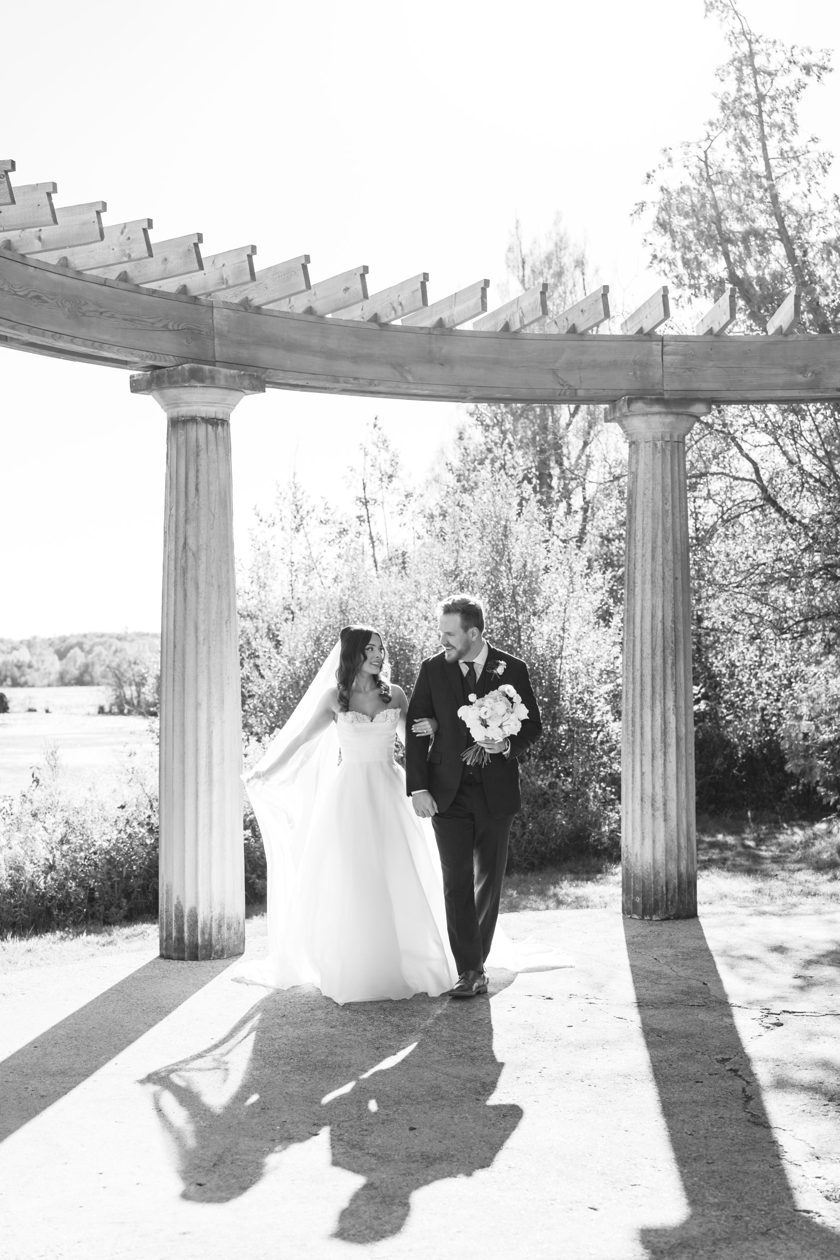 A bride and groom walk hand-in-hand under a pergola with columns, sunlight casting long shadows—a perfect moment for wedding photography. The bride’s flowing gown and veil, and the scenic backdrop, make this a photographer’s dream.