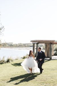 A bride in a flowing white dress and a groom in a dark suit walk together on grass near a lake, smiling, as their wedding photographer captures the moment by a wooden pergola and trees in the background.
