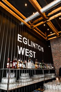 Bottles of liquor and rows of glasses line metal shelves beneath a black wall with large white letters reading “EGLINTON WEST” in a modern Toronto bar, perfect for editorial shoots or wedding celebrations.