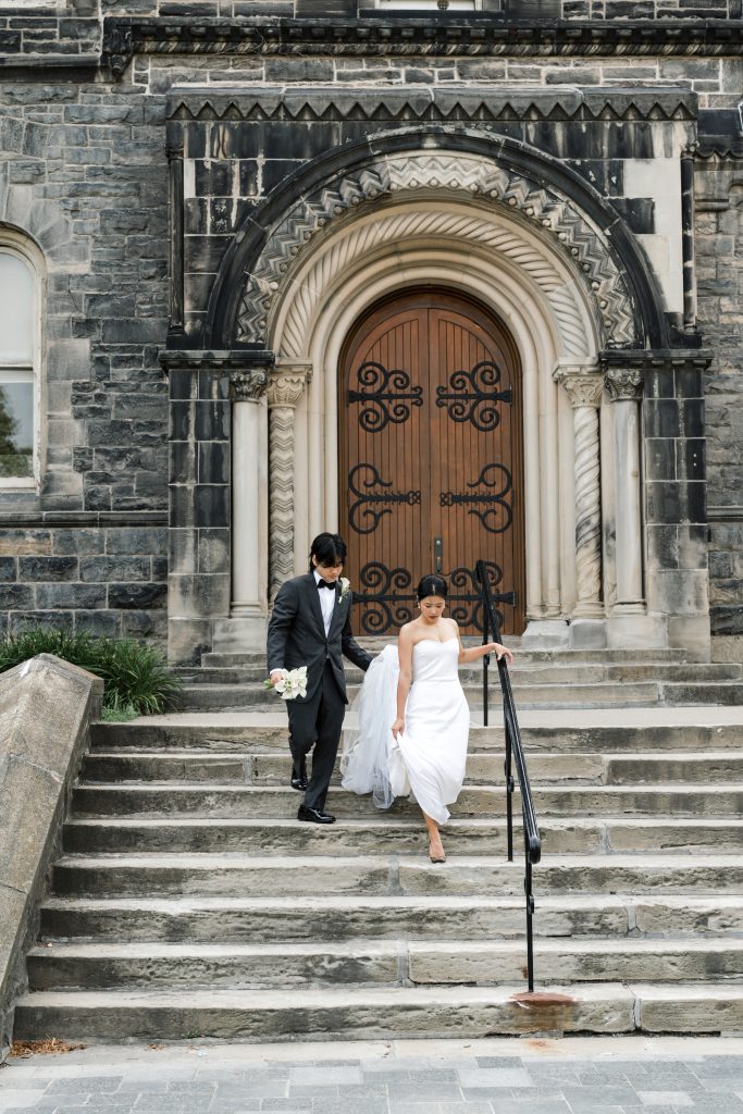 A bride in a white dress and groom in a black suit walk down stone steps outside a historic Toronto building with a large wooden arched door and ornate carvings. This editorial wedding moment captures elegance and city charm.