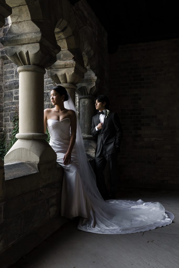 A bride in a strapless white gown and long veil stands by a stone archway, gazing outside, as a photographer captures the moment. The groom, in a dark suit and bow tie, leans nearby in this dimly lit, historic setting.
