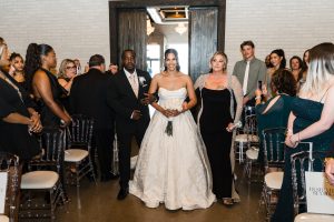 A bride in a white gown walks down the aisle, flanked by two people dressed in black. Guests smile and watch in a modern, indoor wedding venue with white brick walls, as a documentary photographer captures every heartfelt moment.