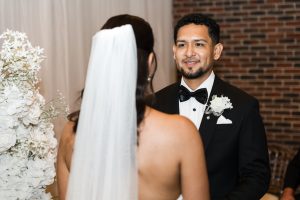 A groom in a black tuxedo and bow tie smiles at a bride in a white dress and veil during their wedding ceremony, beautifully captured by the photographer with white flowers and a brick wall in the background.