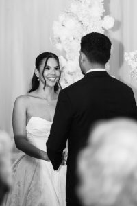 A bride in a strapless gown smiles at the groom as they hold hands during their wedding ceremony, with white floral arrangements in the background, beautifully captured by a Toronto wedding photographer.