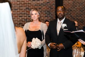 A woman holding white roses and a man in a black suit stand side by side at an indoor wedding ceremony, facing a bride in a white veil. Exposed brick wall in the background, captured in true documentary wedding style by the photographer.