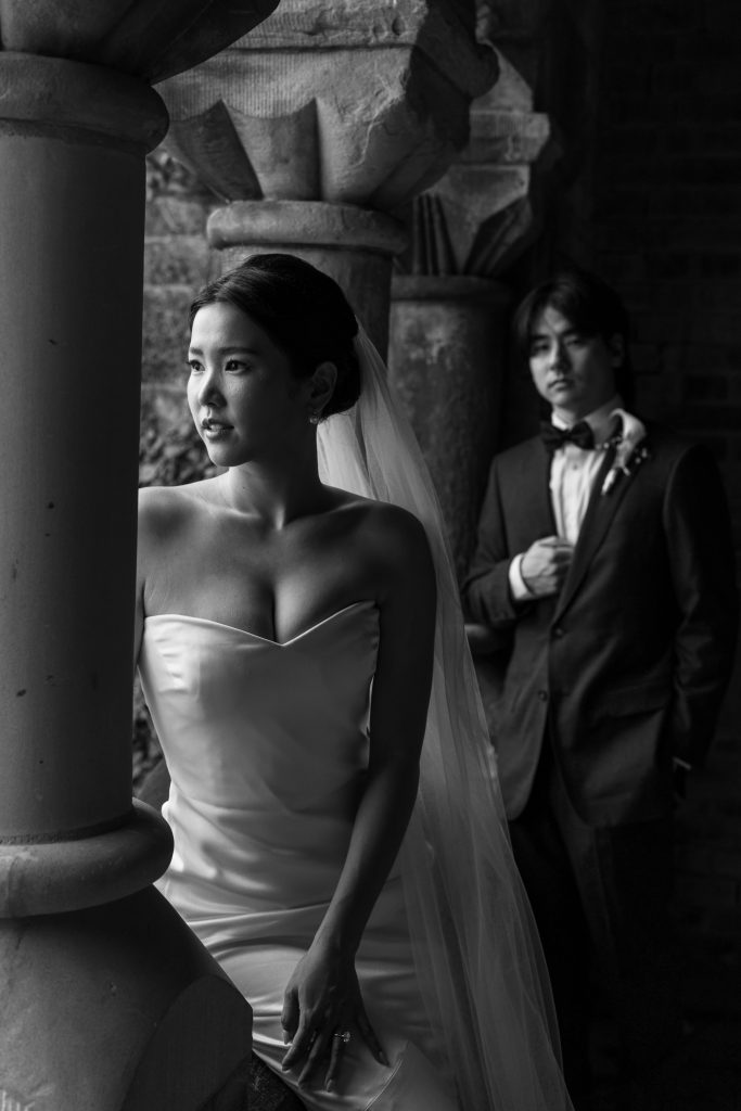 A bride in a strapless gown and veil stands near a stone pillar, gazing outside. In the background, her groom leans against a wall, looking at her. This timeless black and white wedding photography captures a romantic Toronto moment.