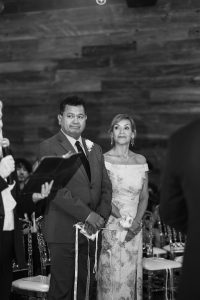 A man in a suit and a woman in an off-the-shoulder floral dress stand side by side, holding a cord, during a formal ceremony. Captured by a documentary photographer, they appear attentive against wooden walls and chairs in Toronto.