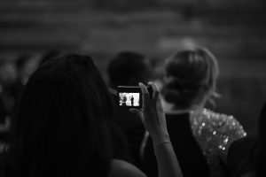A photographer holds up a phone, recording two silhouetted figures on the screen at an indoor Toronto event, surrounded by people in semi-formal attire. The black and white image captures a documentary feel.