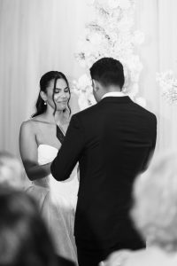A bride in a white dress smiles while holding a card and facing the groom, who is wearing a black suit, during an editorial wedding ceremony with floral decorations in the background.