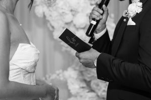A black-and-white documentary photo captures a heartfelt wedding moment: the groom holds a microphone and a booklet labeled Vows, while the bride stands nearby in her strapless dress.