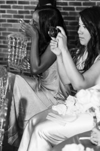 At a Toronto wedding, two women in formal dresses sit on clear chairs. One wipes away a tear, while the other, in an editorial style, holds a camera. Bouquets of white roses rest on their laps against a brick wall backdrop.