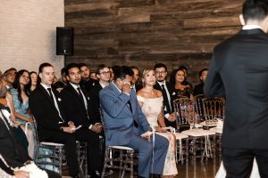A man in a blue suit wipes his eyes while seated among other well-dressed guests at a formal indoor wedding ceremony, with the focus on an individual speaking in the foreground, captured by an editorial photographer.