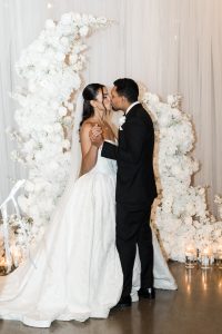 A bride and groom share a kiss while holding hands in front of a white floral arch at their Toronto wedding. Captured by a documentary photographer, the scene glows with lit candles arranged around the flowers.