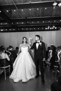 A bride in a strapless gown and a groom in a tuxedo walk hand-in-hand down the aisle, smiling, surrounded by seated wedding guests in a beautifully decorated indoor venue—an elegant moment of editorial wedding photography.