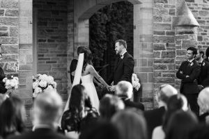 A bride and groom stand holding hands during an outdoor wedding ceremony, facing each other in front of a stone building, with guests seated and attendants nearby, as a photographer captures this editorial moment.