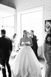 A bride in a white gown smiles and holds a drink at an indoor wedding reception. Captured in black and white, this documentary-style photography highlights guests mingling as sunlight streams through large windows behind them.