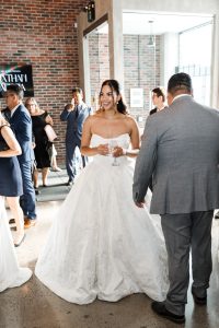 A bride in a strapless white gown holds a glass and smiles while standing among guests in formal attire at a sunlit indoor wedding reception with brick walls, captured in elegant editorial photography.