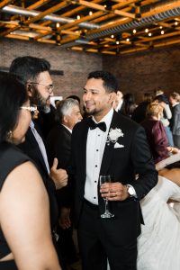 A man in a tuxedo holding a champagne glass smiles as he talks with guests at a lively indoor wedding reception, as a documentary photographer captures candid moments beneath exposed brick walls and glowing string lights.