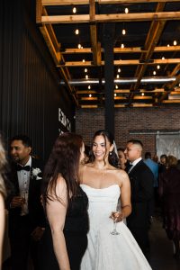 A bride in a white dress smiles and talks with a guest at an indoor Toronto wedding reception, surrounded by well-dressed guests under exposed beams and hanging lights, perfectly captured by the photographer.