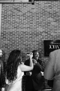 A woman in a white dress feeds cake to a smiling man in a suit at a wedding celebration, surrounded by guests, as a photographer captures the moment in front of a brick wall and a TV screen displaying event text.