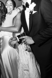 A black and white photo by a Toronto photographer shows a man in a tuxedo holding a champagne glass, while two women in satin dresses embrace and smile in the background, capturing an elegant editorial-style celebration or wedding.
