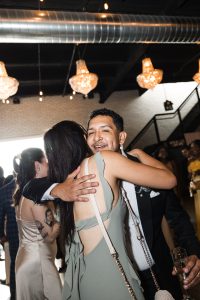 A man in a suit smiles as he hugs a woman in a light green dress at a lively, well-lit indoor event in Toronto. Other guests and chandeliers are visible in the background, giving the scene an authentic, documentary feel.