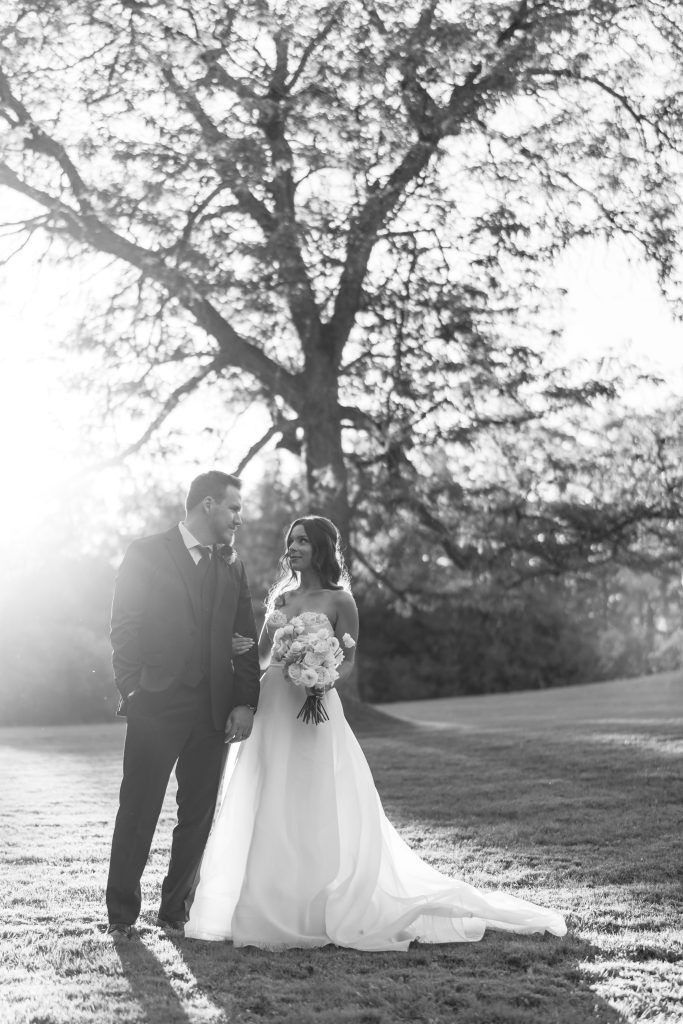 A bride and groom stand together outdoors on grass, holding hands and smiling at each other, with sunlight streaming through a large tree behind them. Captured in black and white, this Toronto wedding photography exudes timeless romance.