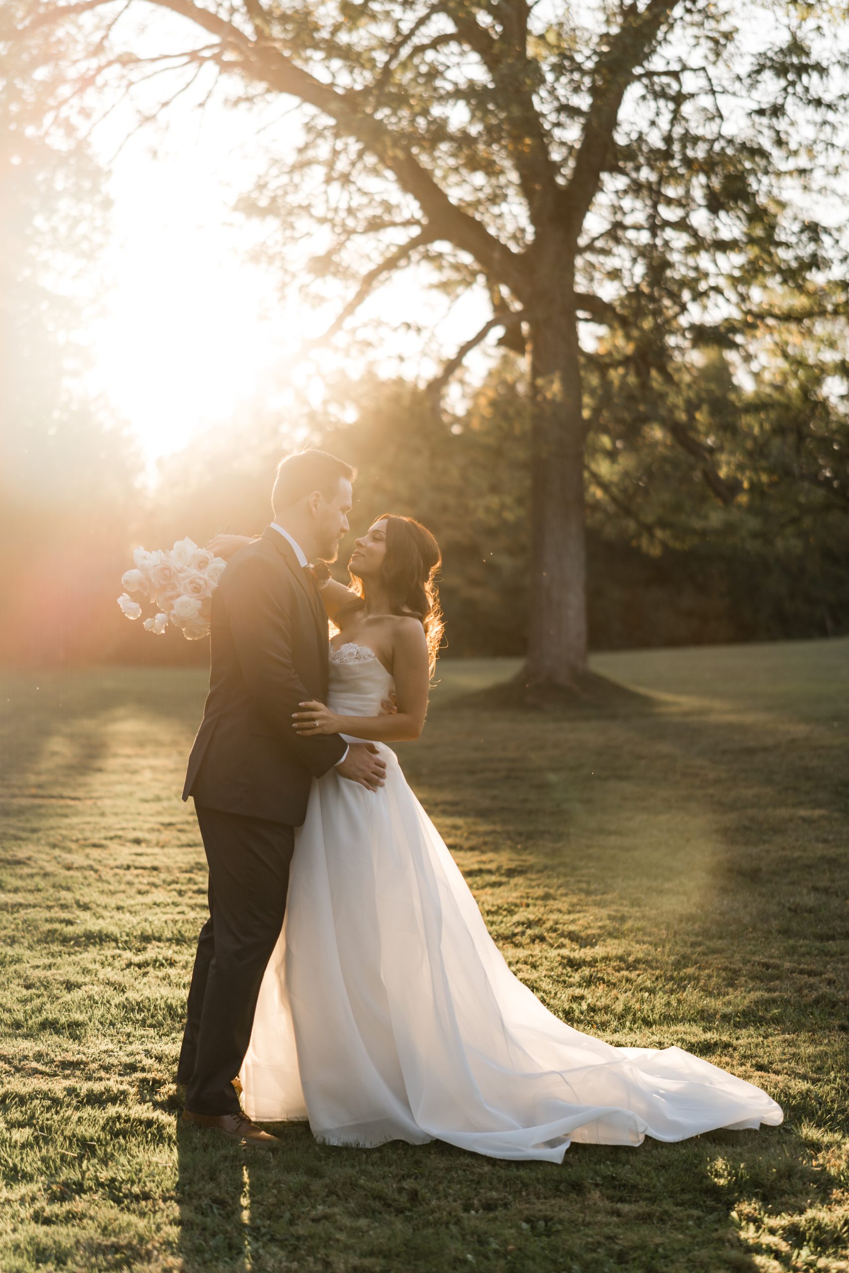 A bride and groom embrace and smile at each other outdoors at sunset during their Toronto wedding, with sunlight streaming through the trees and illuminating the bride’s flowing white dress and bouquet of flowers in a documentary style.