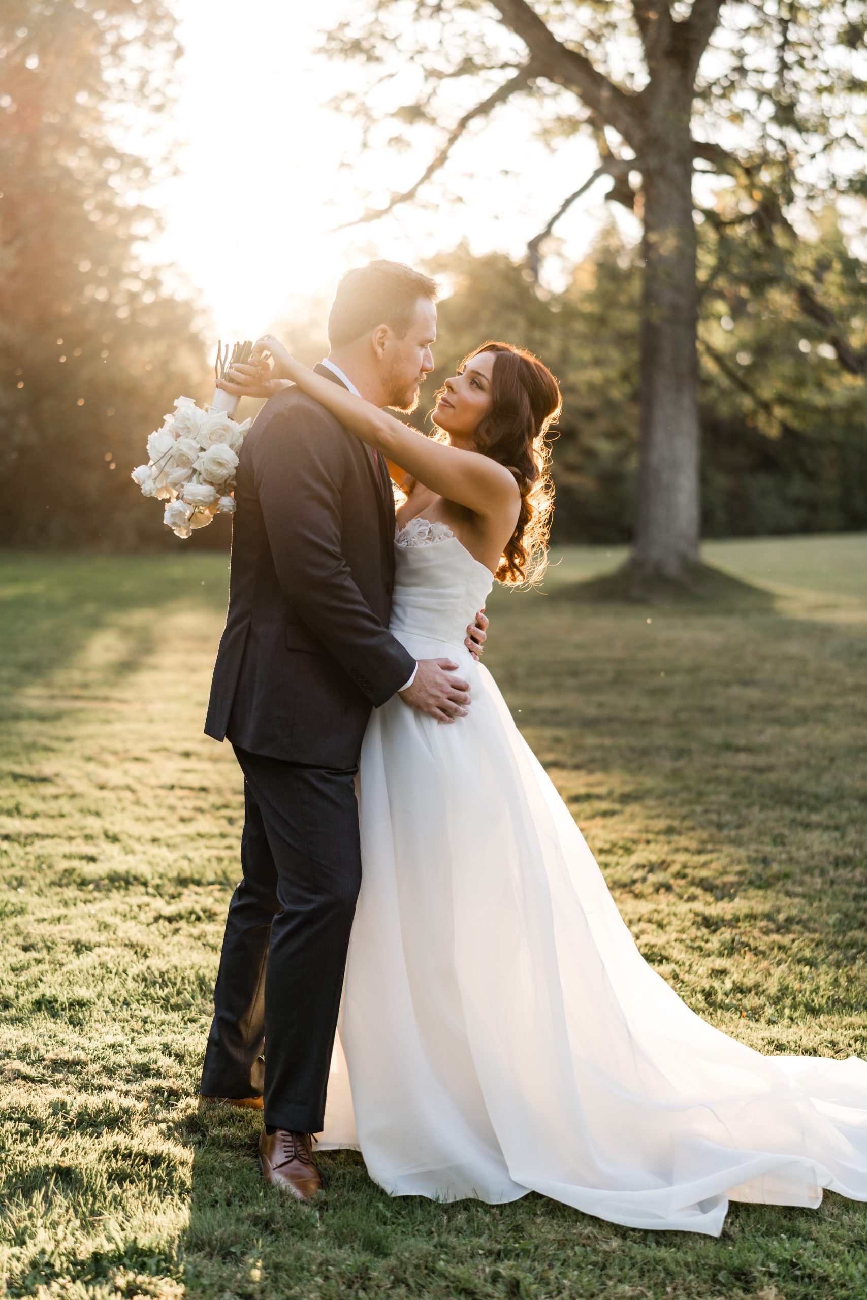 A bride and groom stand outdoors at sunset on a grassy lawn in Toronto, embracing and gazing into each other's eyes in this candid, documentary-style wedding moment. The bride holds a bouquet of white flowers as sunlight streams through the trees behind them.