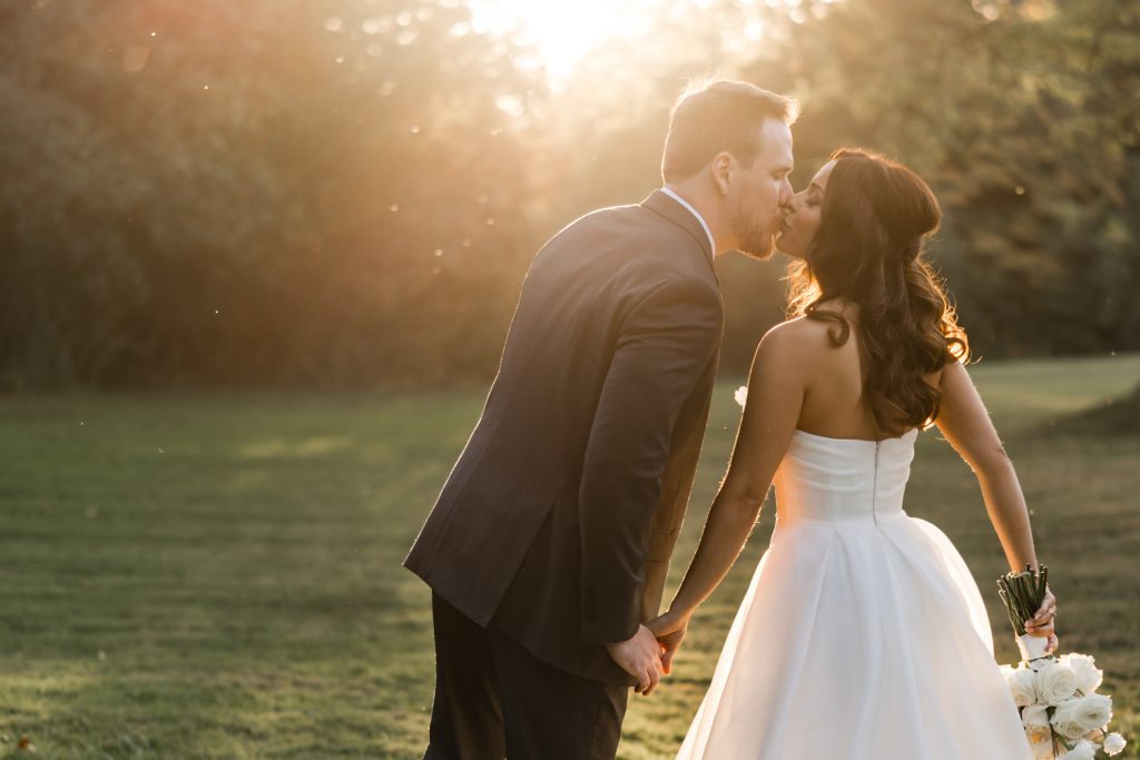 A bride in a white dress and a groom in a suit share a kiss outdoors at sunset, holding hands with a bouquet of white flowers, as their wedding photographer captures the moment amid greenery and soft sunlight.